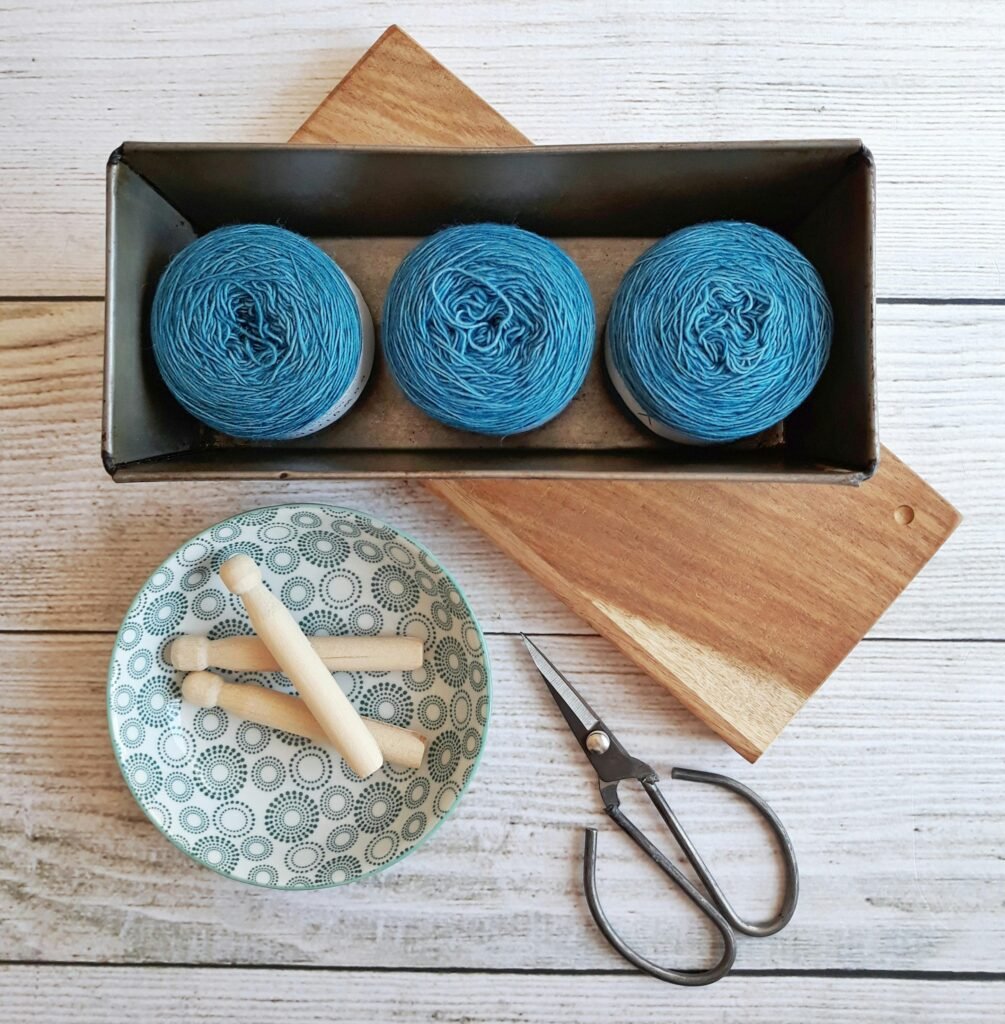 Flat lay of crafting tools, blue yarn, scissors, and spools on wooden table in a rustic setup.